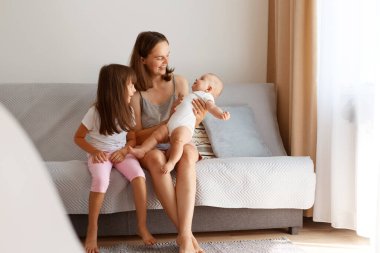 Açık salonda, pencerenin yanındaki koltukta oturan mutlu aile portresi, evde iki kızı olan anne, mutlu insanlar..