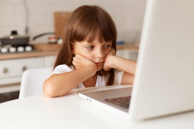 Masada oturan, sıkıcı yüz ifadesiyle defter ekranına bakan, çevrimiçi ders alan, ellerini yüzüne koyan sevimli, esmer kız çocuğunun kapalı çekimleri..