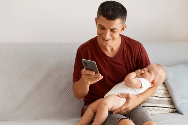 男青年穿着栗色休闲装 坐在房间里灰色沙发上 抱着婴儿 用智能手机 快乐地笑着的室内照片 — 图库照片
