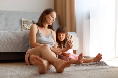 Kolsuz tişört giyen, kısa boylu, kızıyla birlikte yerde oturan, elinde akıllı bir telefon tutan, çocukla hava öpücüğünü kameraya gönderen mutlu bir kadın..