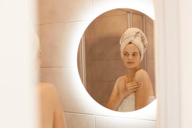 Çekici bir kadının koluna vücut losyonu sürerken, bir kadının ayna yansımasına bakarken, beyaz havluya sarılırken, sabah güzellik prosedürleri yaparken..