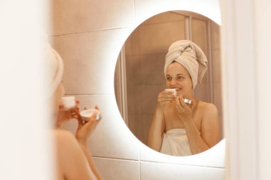 Cildi mükemmel kokulu krem kokan, banyodaki aynadaki yansımasına bakan, omuzları çıplak ve kafasında beyaz havluyla duran çekici bir kadın..