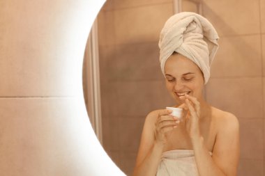 Başında beyaz havluyla gülümseyen kadın başvurmadan önce ellerinde krem tutarak, çıplak omuzlarıyla, olumlu duygular ifade ederek,.