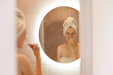 Banyoda poz veren, dişlerini fırçalayan, duş aldıktan sonra hijyen prosedürlerini uygulayan, beyaz saçlı genç bir kadının aynasında yansıması..