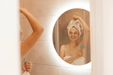 Gülümseyen kadın koltukaltını banyoda tıraş ederken, elinde tıraş makinesi tutarken, aynadaki yansımaya bakarken, beyaz havluya sarınırken, banyoda dikilirken ve tüy döküyorken..