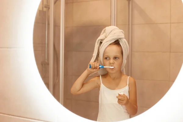 可爱的小女孩站在镜子前刷牙 穿着白色无袖T恤 用毛巾把头发包起来 这是早上的卫生习惯 — 图库照片