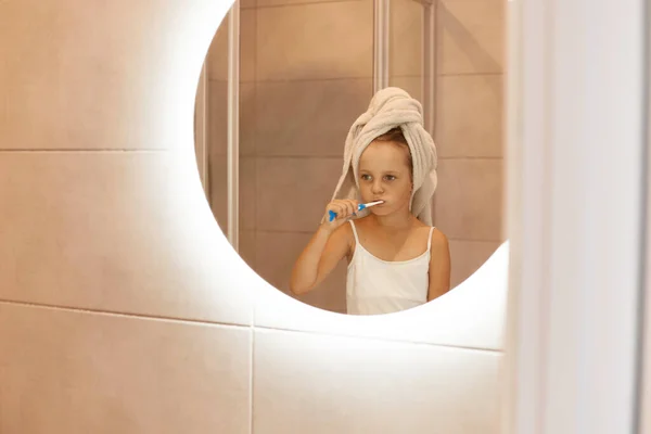 在室内拍摄的女宝宝在浴室刷牙的照片 看着镜子里的自己 穿着白色T恤 用毛巾把头发包起来 卫生习惯 — 图库照片