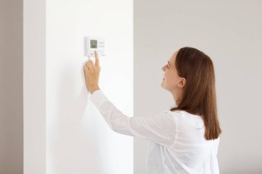 Koyu renk saçlı, beyaz gömlekli, aydınlık odada duran, evde termostatın sıcaklığını ayarlayan güzel bir kadın portresi..