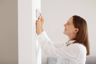 Hoş görünümlü, beyaz bir gömlek giyen, enerji güvenlik ısıtma sistemini izleyen, evdeki dijital kontrollü, termostata dokunan bir kadın..