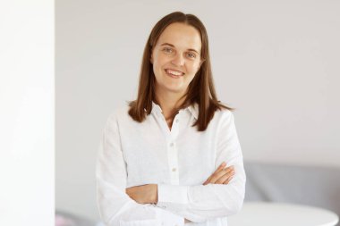 Mutlu, iyimser, hoş görünümlü, beyaz bir gömlek giyen, elleri çapraz duran, özgüvenini ifade eden, kameraya bakan mutlu, iyimser bir kadın..