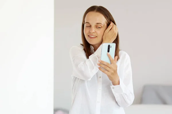 白いカジュアルスタイルのシャツを着た魅力的な幸せな女性の肖像画 手に携帯電話を保持 自撮り スマートフォンのディスプレイを見て 自宅で光の部屋にポーズ — ストック写真