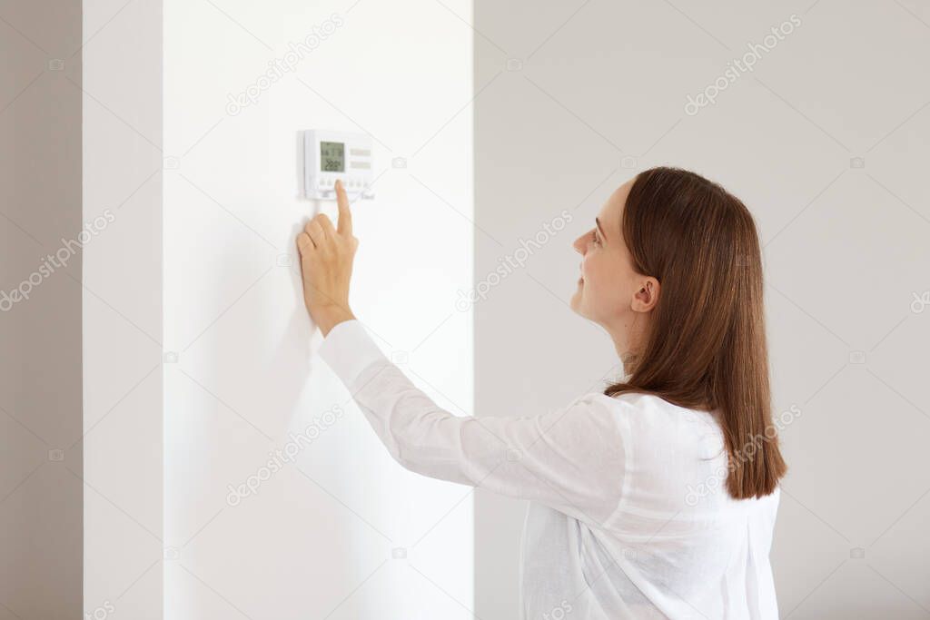 Side view portrait of beautiful female with dark hair wearing white casual style shirt, standing indoor in light room, adjusting central heating temperature at home on thermostat.