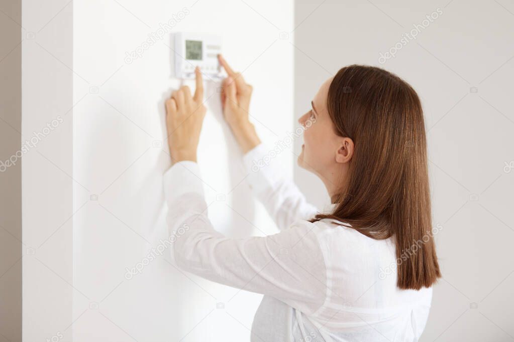 Good looking young adult woman wearing white casual style shirt, using connected appliances device on wall, monitoring energy security heating system, digital control in apartment.
