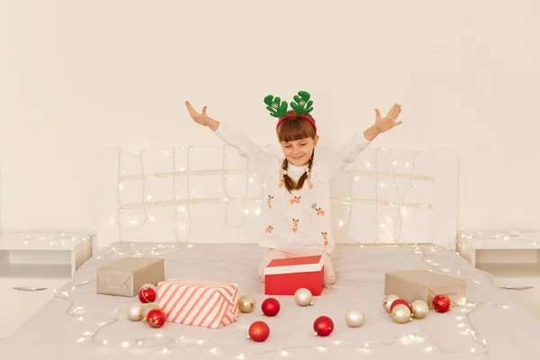 白いセーターとパーティー鹿の角を着て幸せな小さな未就学児の女の子は クリスマスの装飾ボールとベッドの上のオープンプレゼントボックスで遊んでいます 家での季節の休日を祝う — ストック写真