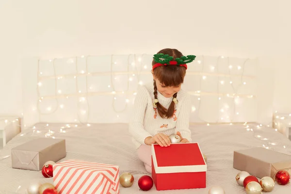 白いセーターとパーティー緑の鹿の角を着て暗い髪の女の子は 新年の装飾 ボールとガーランドとベッドの上に座っている間にクリスマスプレゼントボックスを開く — ストック写真