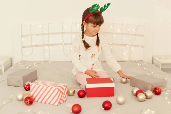 白いセーターとパーティーの緑の鹿の角を着てピグテールを持つ小さな濃縮された女の子はクリスマスボールとガーランドでベッドで遊んで 一人で遊んで 新年を祝いました — ストック写真