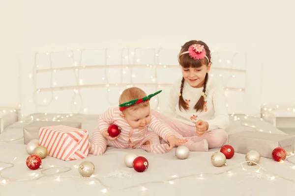 幸せな兄弟の女の子の屋内ショットクリスマスの装飾でベッドの上に座って ボールで遊んで 一緒に新年を祝う カジュアルな服を着て — ストック写真
