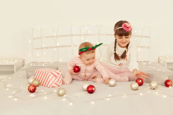 魅力的な姉妹の肖像画 ライトで飾られたベッドの上でクリスマスボールで遊んでかわいい女の子 カジュアルな服装をした子供たち 幸せな新年 — ストック写真