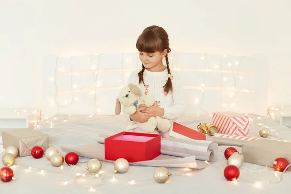白いセーターを着たチャーミングな女の子は柔らかいおもちゃの犬を受け入れ クリスマスの装飾とガーランドでベッドに座って 彼女の現在を見て 新年の前夜を祝う — ストック写真
