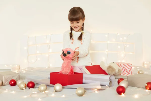 柔らかいおもちゃを保持白いセーターを着て魅力的な少女の屋内ショット 笑顔で彼女の新年の贈り物を見て クリスマスの装飾とガーランドとベッドの上に座って — ストック写真