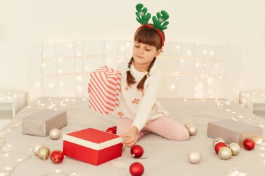 Yoğunlaşmış, siyah saçlı, beyaz kazak giyen, geyik boynuzları takan, karnaval çemberi takan, kış tatiline hazırlanan, Noel hediyelerini paketleyen küçük bir kız..