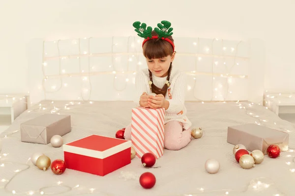 白いセーターと鹿の角カーニバルの輪を身に着けているかなり小さな女の子の屋内ショット クリスマスプレゼントとベッドの上に座って 手でXマスツリーの装飾のためのボールを保持 — ストック写真