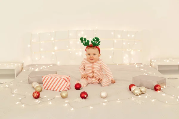 魅力的なかわいい幼児の女の子はベッドと鹿の角カーニバルの輪を着てベッドの上に座ってカメラを見て 子供の生活の中で興味 最初のクリスマスと新年を表現 — ストック写真