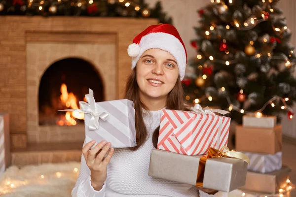 白いセーターとサンタクラスの帽子を身に着けている肯定的な楽観的な女性の肖像画は 暖炉とクリスマスツリーとお祝いの部屋にいること 新年のプレゼントを保持 — ストック写真