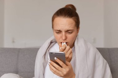 Kapalı alanda, battaniyeye sarılı koltukta oturan, grip belirtileri gösteren, akıllı telefon kullanan, internetten tedavi hakkında bir şeyler okuyan hasta bir kadın.,
