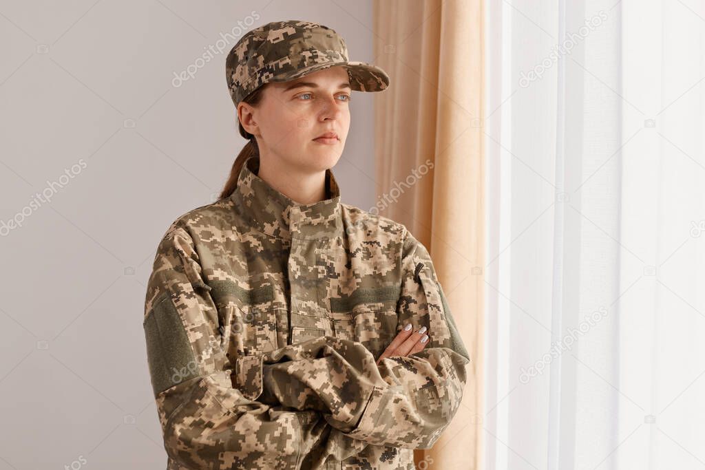 Indoor shot of serious woman soldier returning home after army, posing at home in living room, standing with crossed arms and looking away with serious expression.