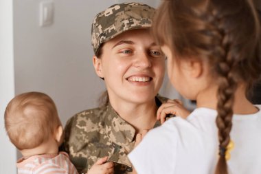 Kamuflaj üniforması ve şapka giymiş gülümseyen iyimser asker annenin yakın plan portresi, askeriyeden sonra eve döndüğünde kızlarıyla buluşması..