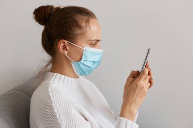 Beyaz kazak ve tıbbi maske giyen, elinde cep telefonu olan, alet görüntüsüne bakan, e-postaları kontrol eden, gribi tedavi etmek için bilgi arayan sağlıksız kadınların yan görüntüsü..