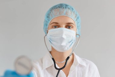 Çekici beyaz kadın doktor portresi tıbbi üniforma, başlık ve cerrahi maske takıyor, steteskopu kameraya gösteriyor, sakin bir yüz ifadesi takınıyor..