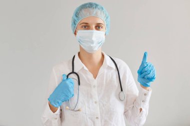 Tıbbi üniforma, başlık ve cerrahi maske takan, ayakta duran, kameraya bakan, baş parmağını kaldıran, virüs tedavisinde yeni yöntemleri onaylayan pozitif kadın doktorların kapalı çekimleri..