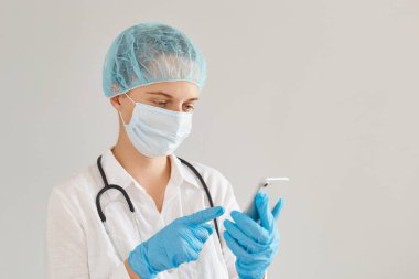 Tıbbi üniforma, şapka ve cerrahi maske takan genç yetişkin kadın doktor portresi elinde cep telefonuyla duruyor, e-postaları kontrol ediyor ya da bilgi arıyor..