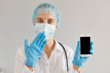 Tıp şapkası, eldiven ve koruyucu maske takan, reklam veya promosyon için telefonu boş gösteren, ağzı eliyle kapatan şaşırmış kadın doktor..
