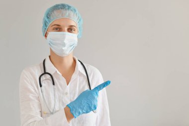 Tıbbi başlık, eldiven ve koruyucu maske takan iyimser kadın doktor portresi işaret parmağıyla kenarda duruyor, reklam için fotokopi alanı sunuyor..