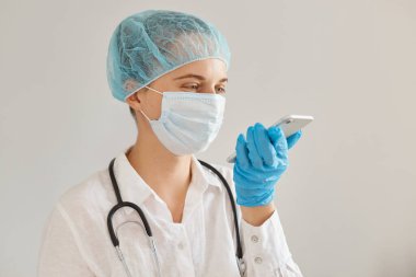 Tıbbi başlık takan, eldiven takan, koruyucu maske takan, cep telefonu kullanan, sesli mesaj kaydeden, olumlu duygu ve mutluluğu ifade eden kadın doktorların kapalı çekimleri..