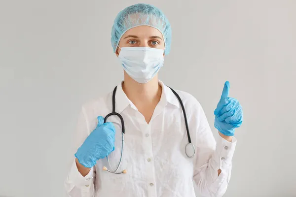 穿着医疗制服 头罩和外科口罩的女医生站在室内拍照 看着相机 竖起大拇指 批准了新的病毒治疗方法 — 图库照片
