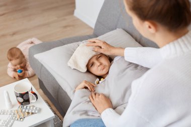 Alnında ıslak havluyla kanepeye uzanmış hasta küçük bir kızın üst görüntüsü, yüksek ateşten muzdarip, annesi sağlıksız kızına bakıyor, soğuk havadan grip olmuş..