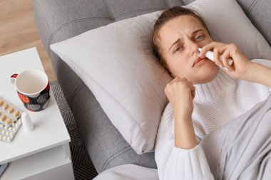 Kanepede yatıp burun spreyi kullanan, kameraya bakan, Grippe 'i tedavi eden, grip belirtileri gösteren sağlıksız hasta bir kadın tedavi edilmeli..