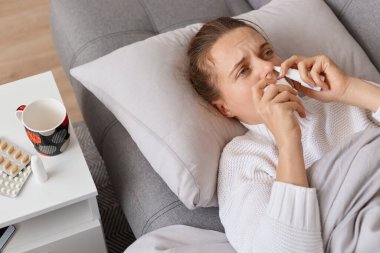 Kanepede yüksek ateşle yatan, nezle ve nezle olan, burun spreyi kullanan üzgün duyguları ifade eden, gribe yakalanan hasta bir kadının portresi..
