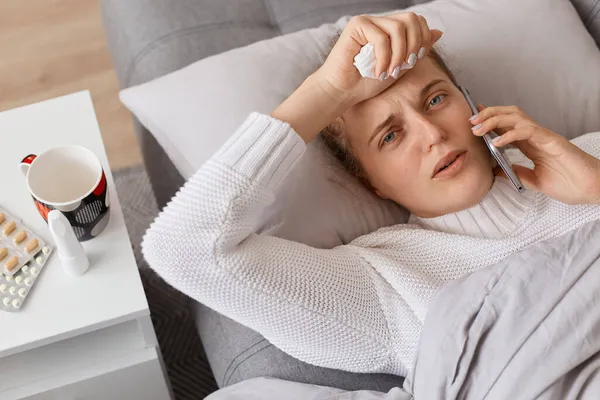 Indoor shot of sick girl lying in bed and uses smartphone to talk to her doctor, suffering headache and high temperature, looking at camera, expressing sorrow.