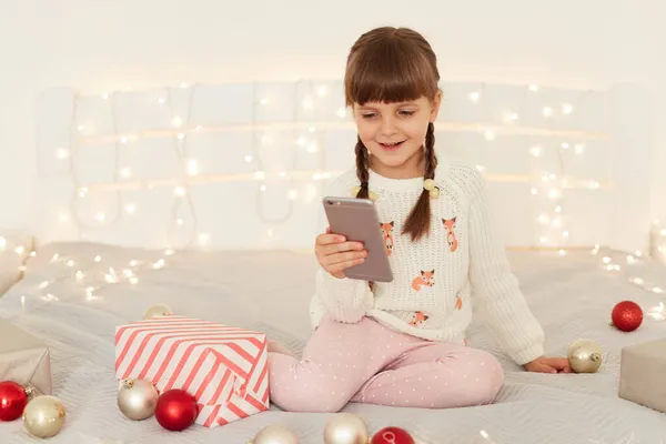 幸せな暗い髪の女性の子供の屋内ショットピグテール手に携帯電話を保持し 自撮りを取ります スマートフォンのカメラを見て 喜んで笑顔 クリスマスを祝います — ストック写真