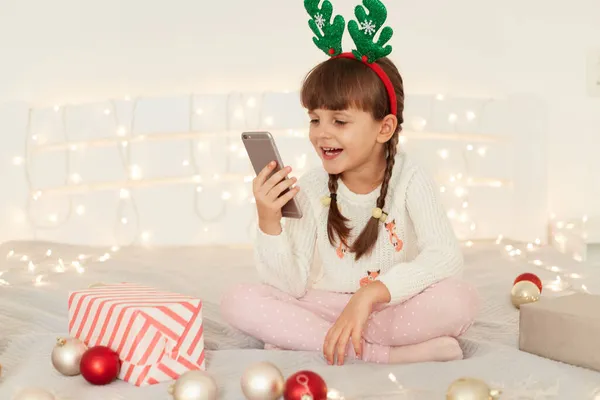 幸せな正の暗い髪の女性の子供とともにピグテールを手に携帯電話を持っています 小さな女の子は スマートフォンの画面を見て 幸福を表現します ビデオ通話を持っています — ストック写真