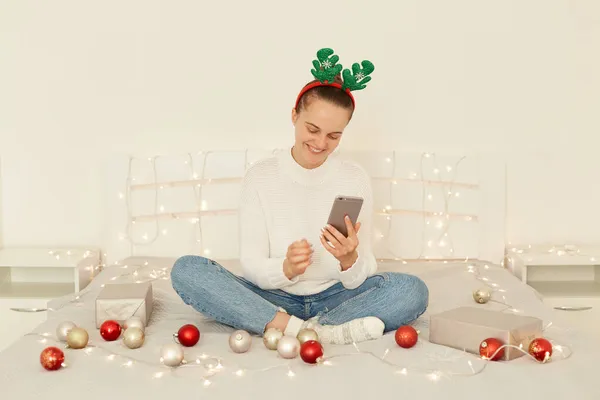 白いセーターのジーンズを着た魅力的な笑顔白人女性 パーティー小道具フープ ベッドルームでベッドに座って オンライン通信のための携帯電話を使用して クリスマスの挨拶 — ストック写真