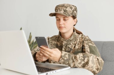 Kamuflaj üniforması ve şapka giyen, evde poz veren, taşınabilir bilgisayarın önünde oturan ve cep telefonu kullanan sakin kadın askerin yatay görüntüsü sakin bir ifadeye sahip..