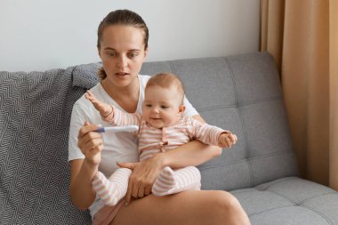 Beyaz tişörtlü, küçük kızıyla koltukta oturan ve hamilelik testini elinde tutan şaşırmış bir kadın pozitif sonuçtan hayrete düşüyor..