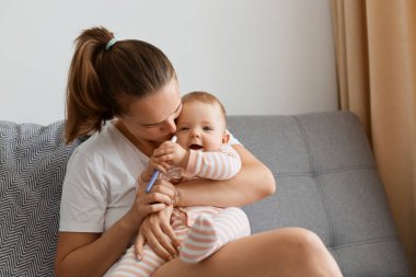 Beyaz tişörtlü, siyah saçlı bir kadının küçük kızıyla kanepeye oturup, elinde hamilelik testi tutarken ve büyüleyici çocuğunu öperken çekilmiş bir fotoğraf..