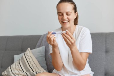 Elinde hamilelik testi olan günlük kıyafet giyen, beyaz saçlı, gülümseyen kadın portresi, oturma odasında otururken poz vermek, pozitif sonuç almak,.